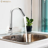A silver kitchen sink tap with water flowing into a stainless steel sink, positioned against a white countertop and a wall with a plant decoration.
