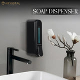 Black soap dispenser mounted on a wall next to a sink & tap with a vase of flowers.