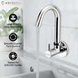 An image of a single-hole wall-mounted chrome mixer tap with a smooth, curving spout, actively dispensing water with its features on the left side on a faded white background.