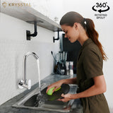 A woman is using a silver wall-mounted kitchen sink tap with a rotating spout over a sink, washing a green dish.