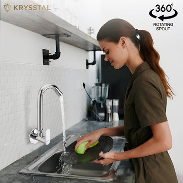 A woman is using a silver wall-mounted kitchen sink tap with a rotating spout over a sink, washing a green dish.