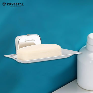 A square Silver soap dish holder, carrying soap, mounted on blue wall with white shampoo bottle on right.