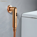 Rose gold colored health faucet with a shower tube and wall hook beside white toilet.