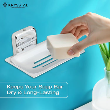 A silver square soap dish holder, carrying a hand-held soap, mounted on a blue wall with a written tagline at the bottom & beside a white pot with a plant.