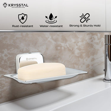 A wall-mounted square silver soap dish holding soap, reflecting on the holder, fitted on a brown textured wall, with its features on top & a white washbasin with tap at the bottom.