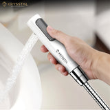 Handheld shower head with water flowing, held by a hand, on a blurred background with Krysstal branding.